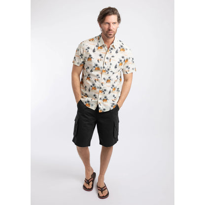 Front view of a man wearing a Weird Fish short sleeve Faraway shirt in Ecru with a tropical island, palm trees, sun and sailboat pattern with navy shorts and flip flops.