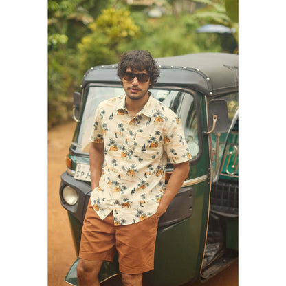 Front view of a man wearing a Weird Fish short sleeve Faraway shirt in Ecru with a tropical island, palm trees, sun and sailboat pattern with orange shorts outdoors.