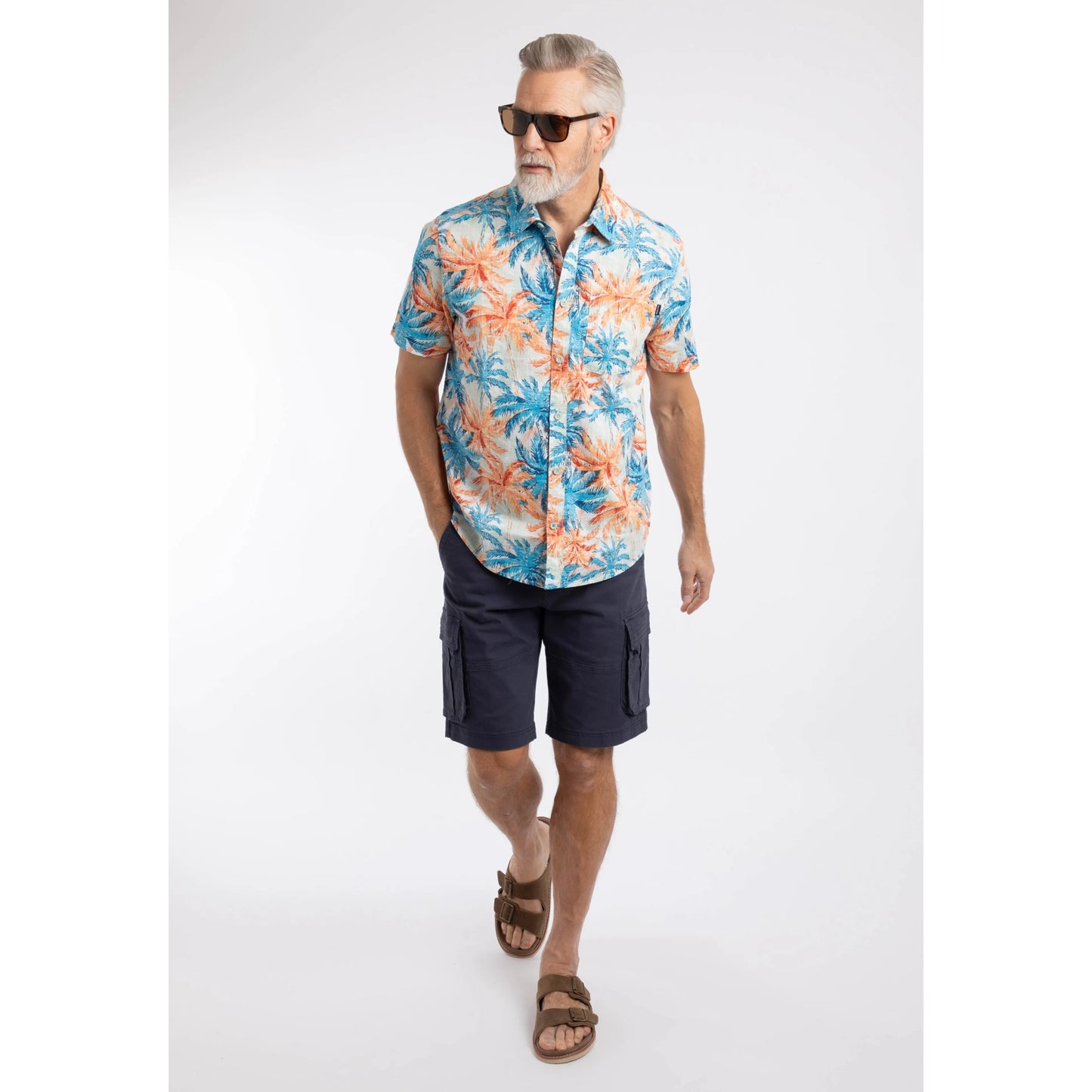 Man wearing a Weird Fish short sleeve Faraway shirt in Paprika orange and blue with a tropical palm tree pattern, with navy shorts and brown sandals