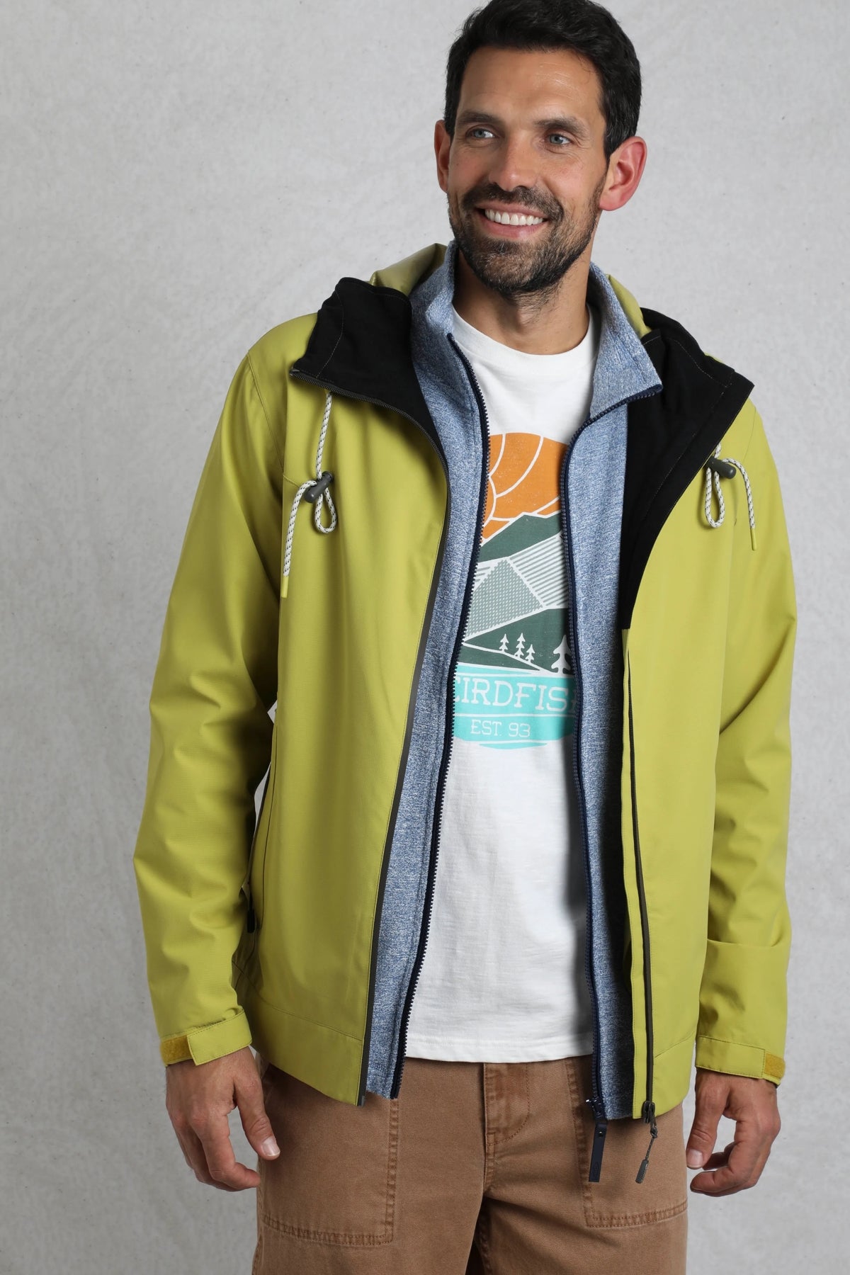 Weird Fish mens waterproof jacket tarragon in green worn by man with brown pants and graphic tee