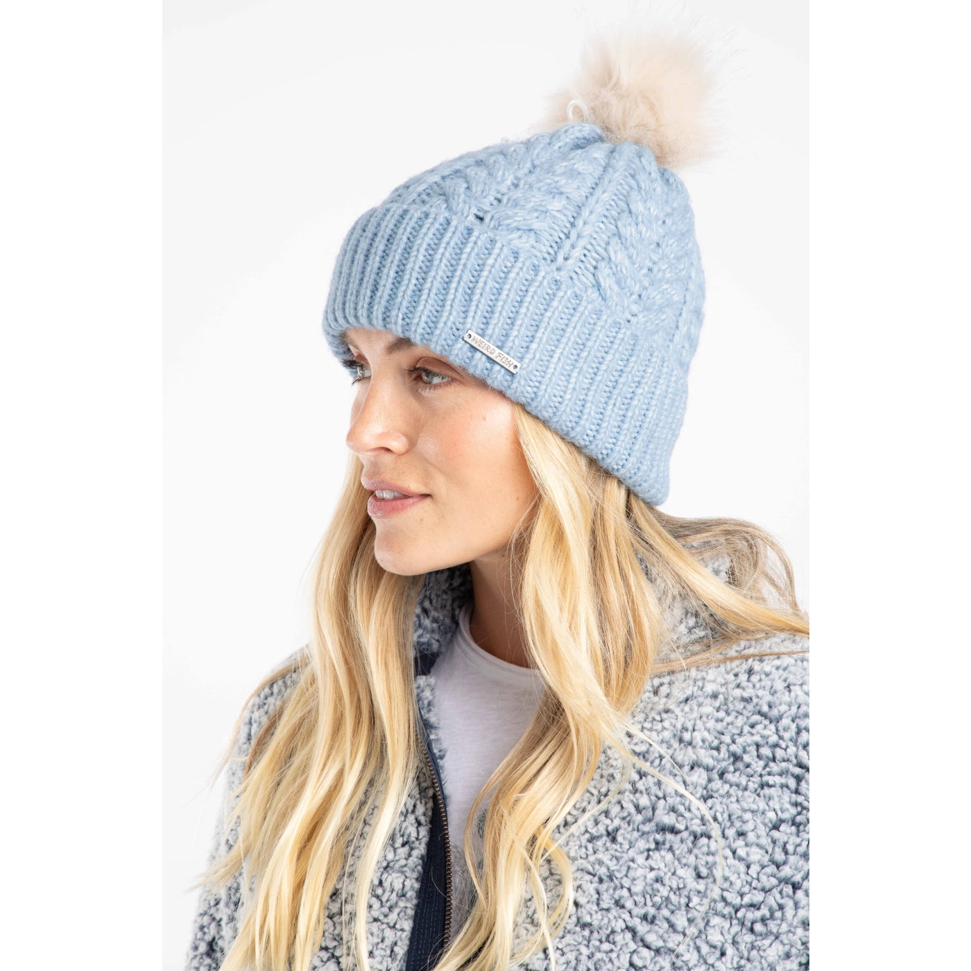 Woman wearing a Weird Fish Lake Blue Allerton cable knit beanie with a faux fur pom-pom on a white background