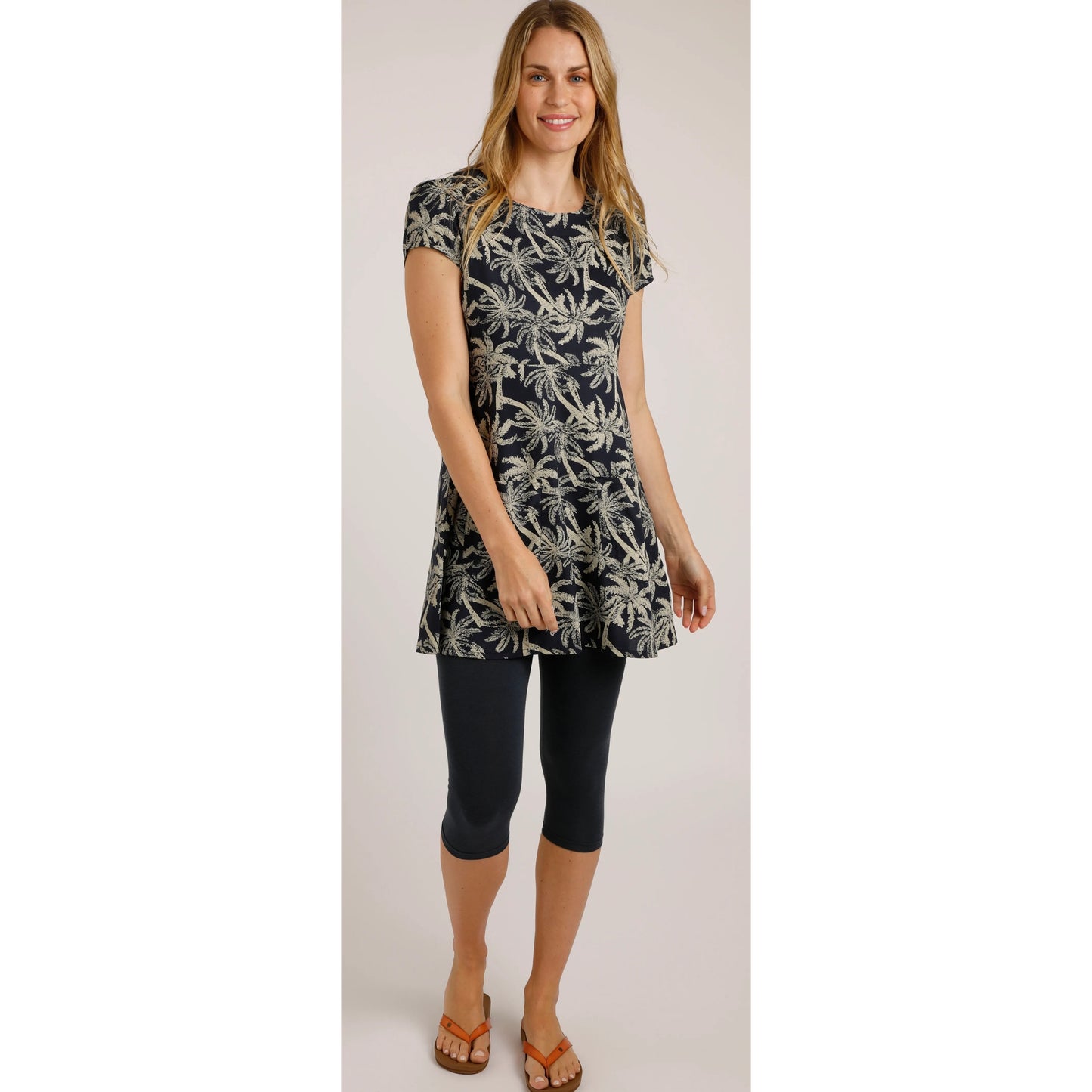 A woman wearing a Weird Fish short sleeve Breeza tunic in Midnight Navy with a beige palm tree pattern with navy leggings and flip fops