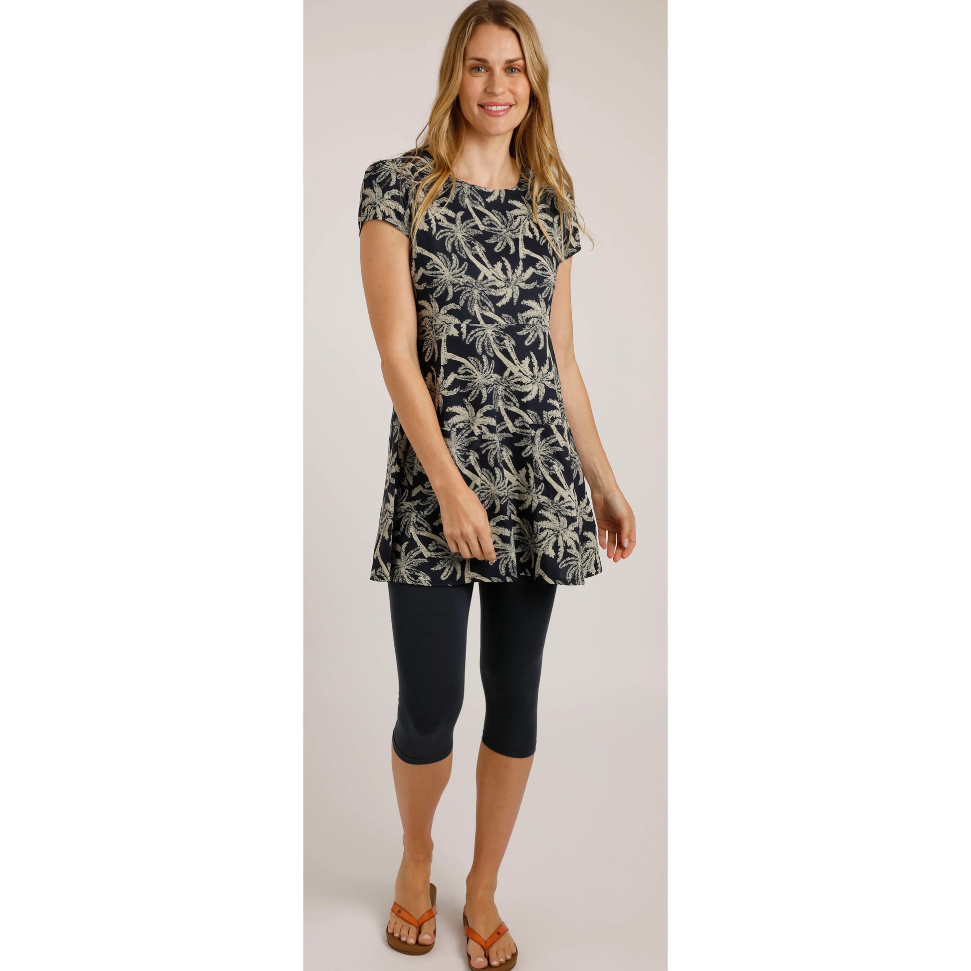 A woman wearing a Weird Fish short sleeve Breeza tunic in Midnight Navy with a beige palm tree pattern with navy leggings and flip fops
