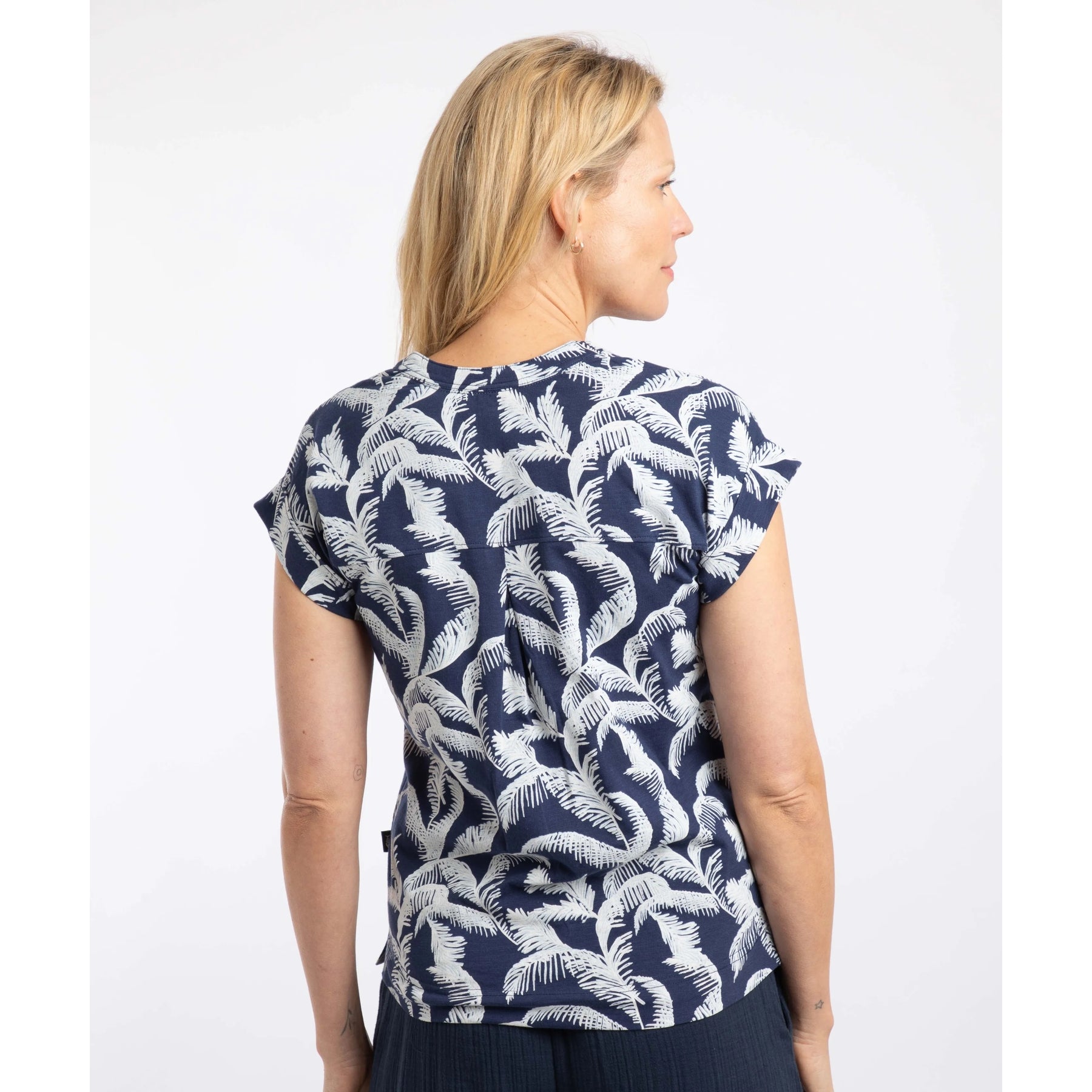 Back view of a woman wearing a short sleeve Weird Fish Alia t-shirt in Ink blue with an ecru palm tree leaf print on a white background