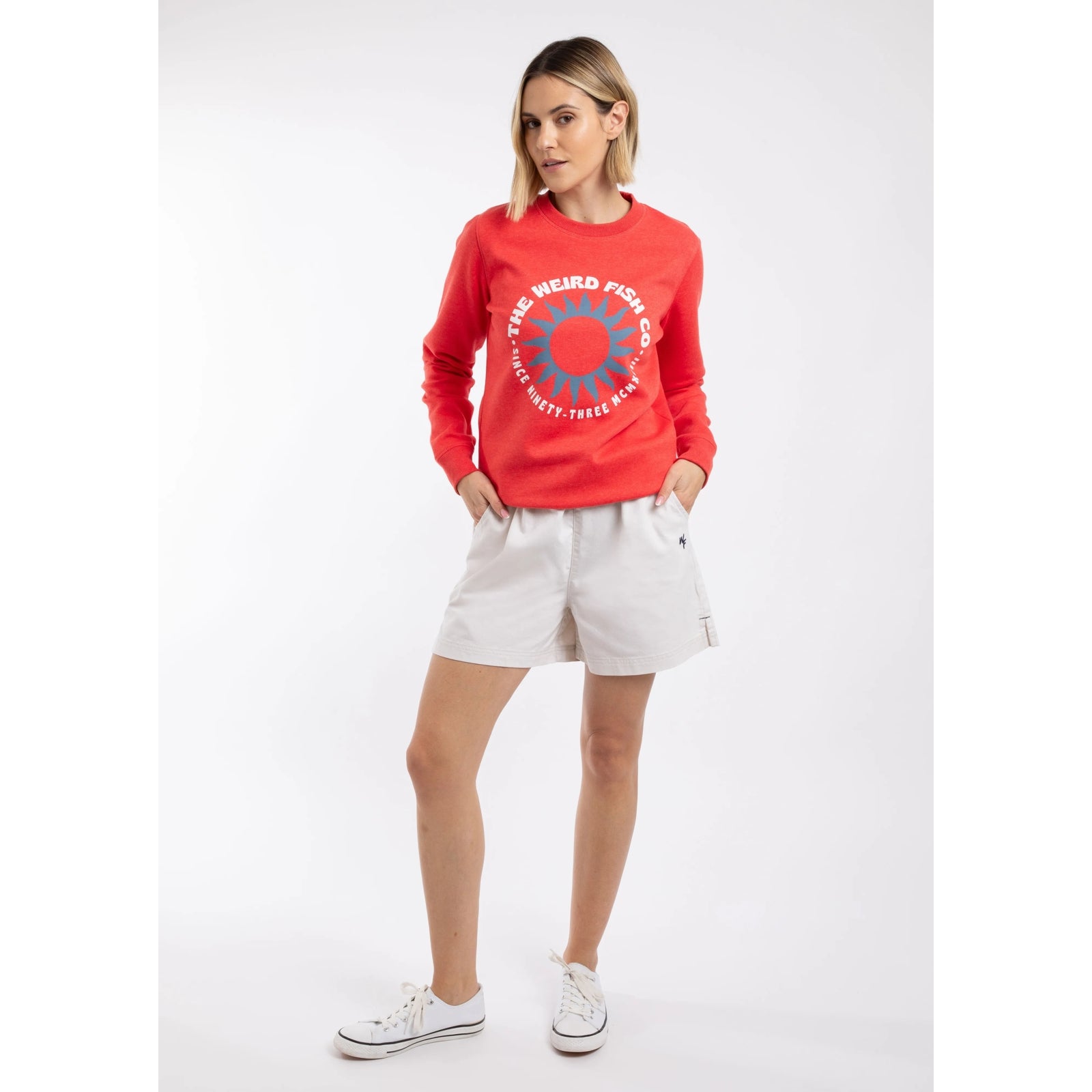 A woman wearing a Weird Fish Aurora crew neck sweatshirt in a bright coral colour with bold circular logo on the front with white shorts.