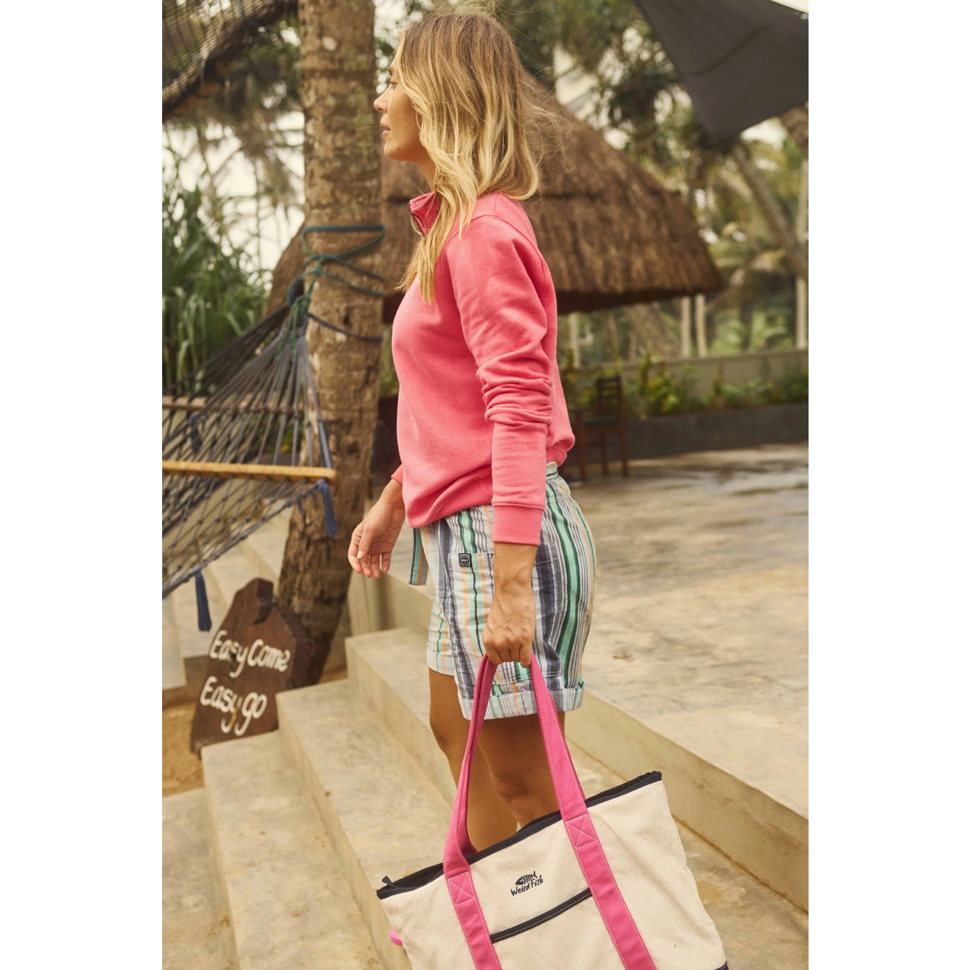 Side view of a woman wearing a Weird Fish Avery 1/4 zip plain sweatshirt in Flamingo Pink with mutlticolour strip shorts