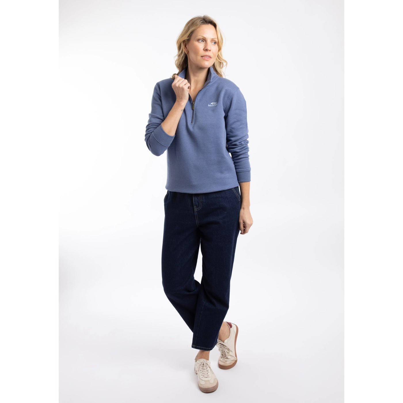 Woman wearing a Weird Fish Avery 1/4 zip plain sweatshirt in Faded Blue with black trousers and beige trainers