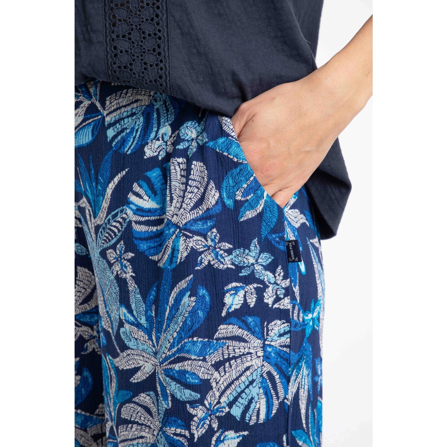 Close-up view of a woman wearing a pair of Weird Fish Sarita cropped trousers in indigo blue with tropical leaf pattern, with one hand in a hip pocket and a small logo label sewn near the pocket.