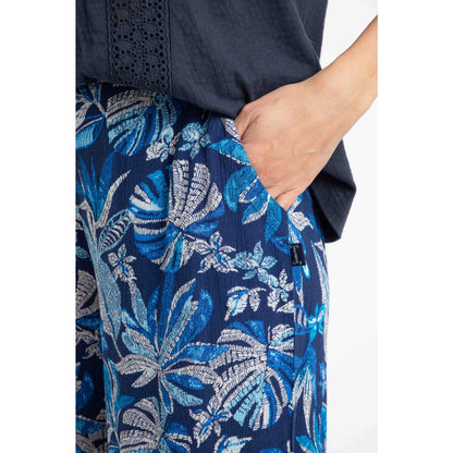 Close-up view of a woman wearing a pair of Weird Fish Sarita cropped trousers in indigo blue with tropical leaf pattern, with one hand in a hip pocket and a small logo label sewn near the pocket.