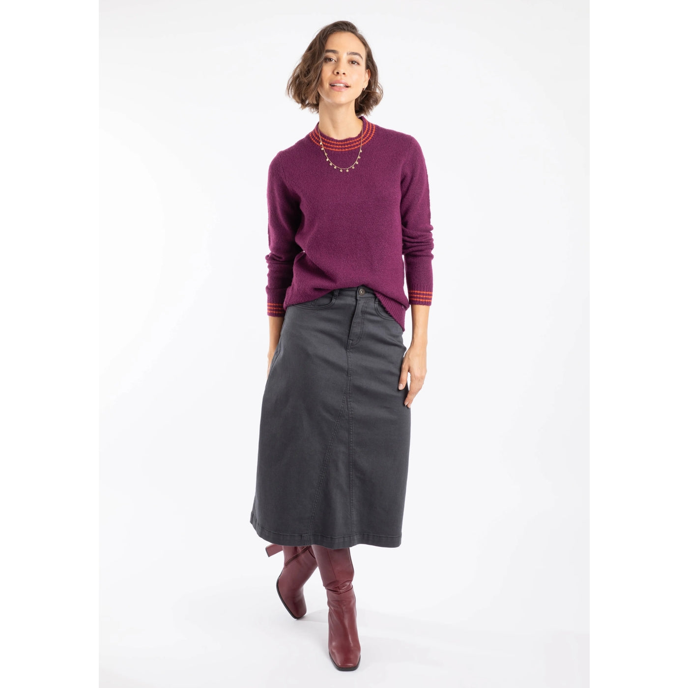 Woman wearing a Weird Fish Beet Purple knitted Dawkins jumper with a grey skirt and brown boots on a white background.
