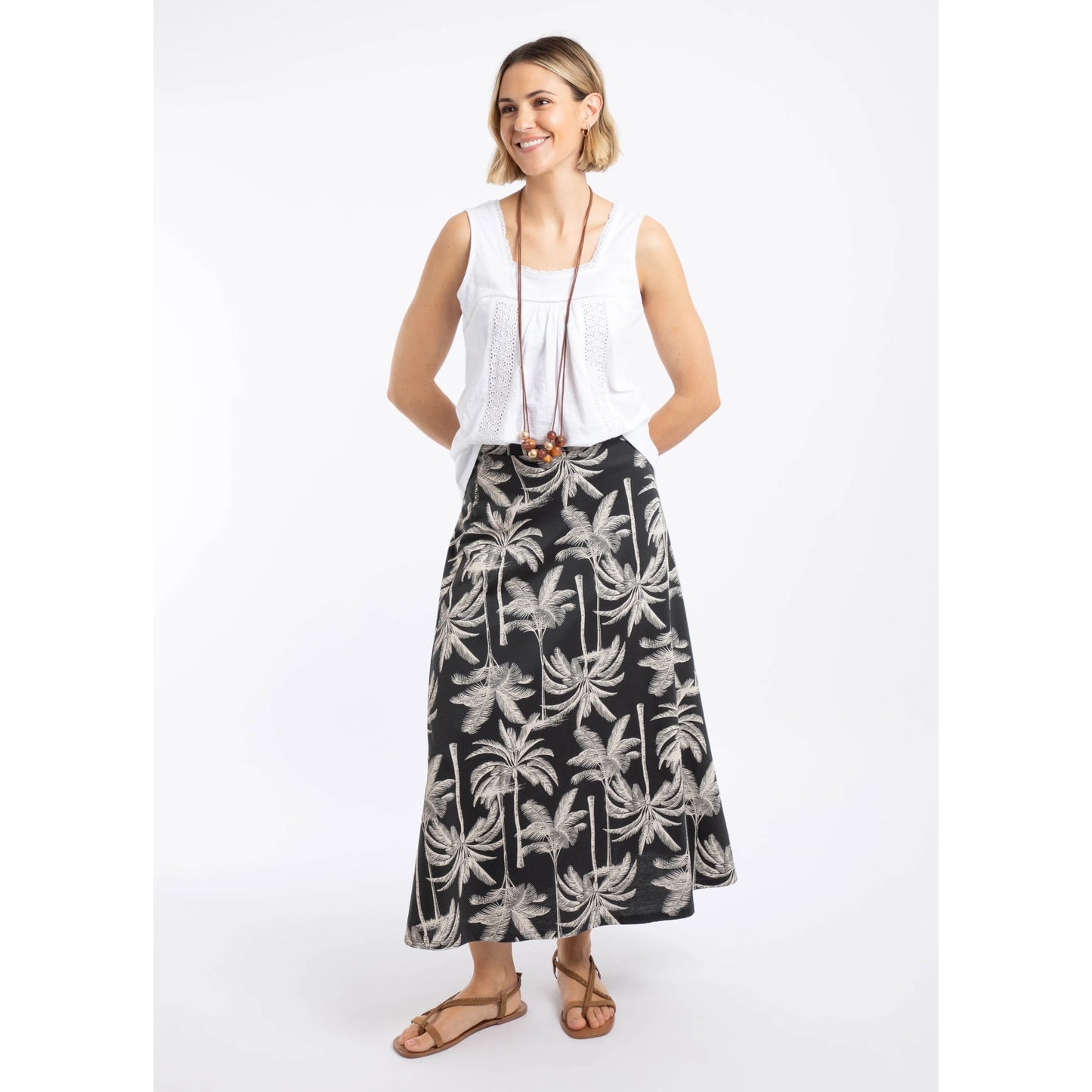 A woman wearing a Weird Fish Eloriah midi jersey skirt in black with a beige palm tree pattern with a white vest and brown sandals.