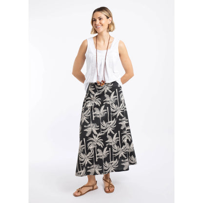 A woman wearing a Weird Fish Eloriah midi jersey skirt in black with a beige palm tree pattern with a white vest and brown sandals.