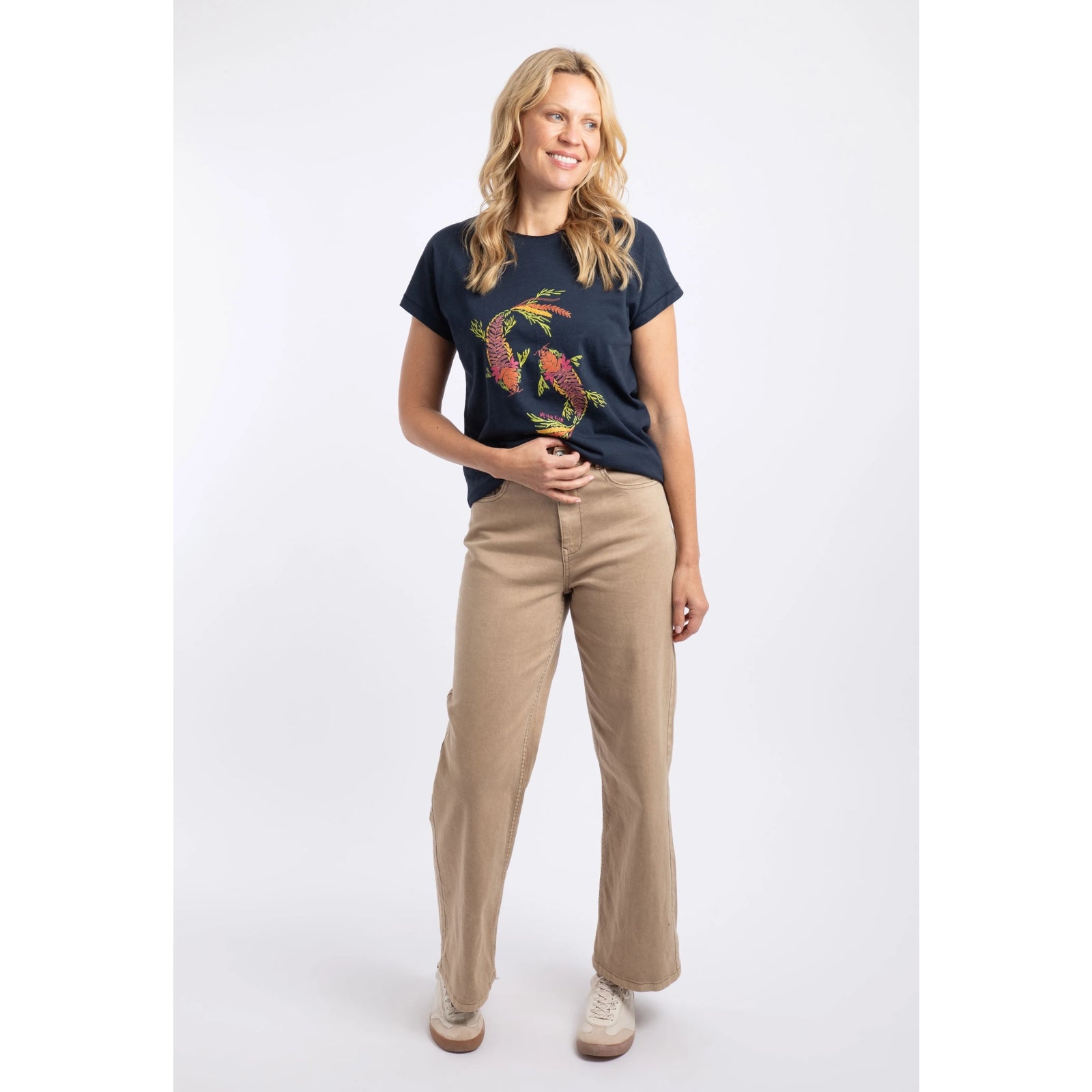 A woman wearing a Weird Fish short sleeve navy t-shirt featuring an abstract koi carp print made up of fern leaves on the front with beige trousers and white trainers.
