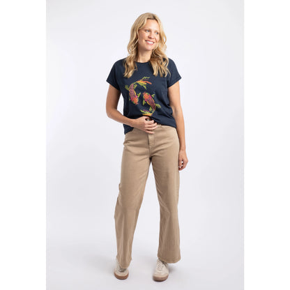 A woman wearing a Weird Fish short sleeve navy t-shirt featuring an abstract koi carp print made up of fern leaves on the front with beige trousers and white trainers.