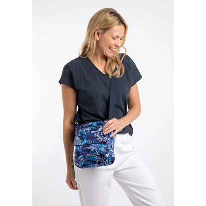 Woman wearing a Weird Fish Kait cross body bag in navy with a blue tropical leaf print, adjustable shoulder strap and front pocket.