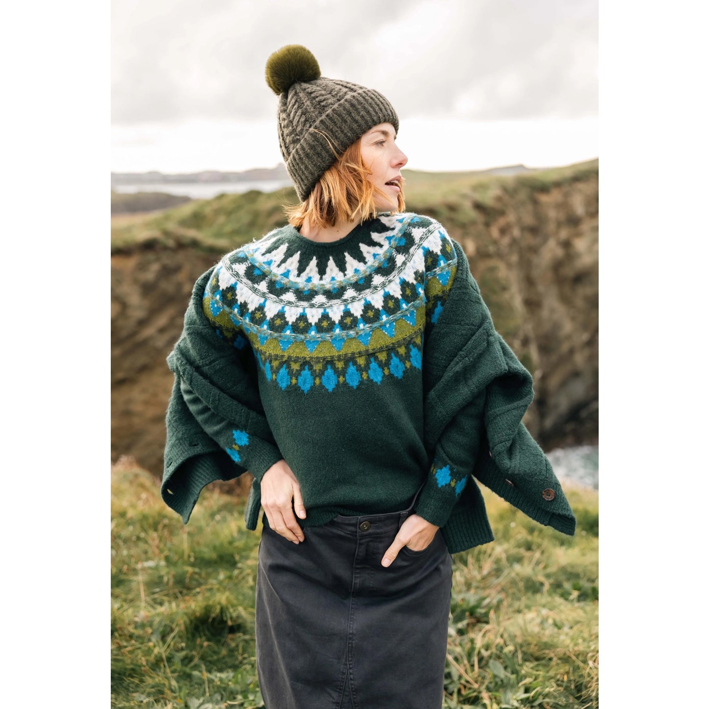 Front view of a woman wearing a Weird Fish knitted Lowell jumper in Bottle Green, with a Fair Isle pattern across the chest and shoulders, stood outdoors wearing a bobble hat and jacket.