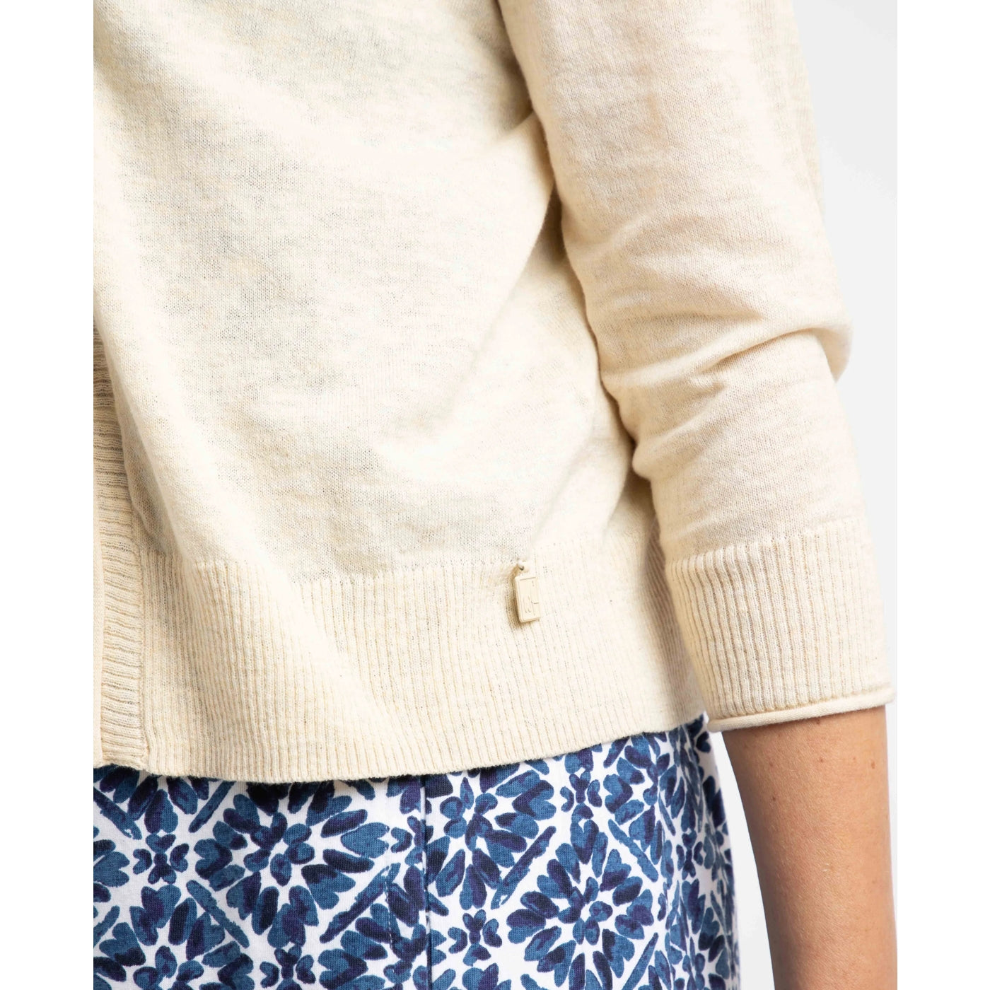 Close-up view of the logo fob and ribbed sleeves and hem of a women's Weird Fish Limon 3/4 sleeve shrug cardigan in Light Cream
