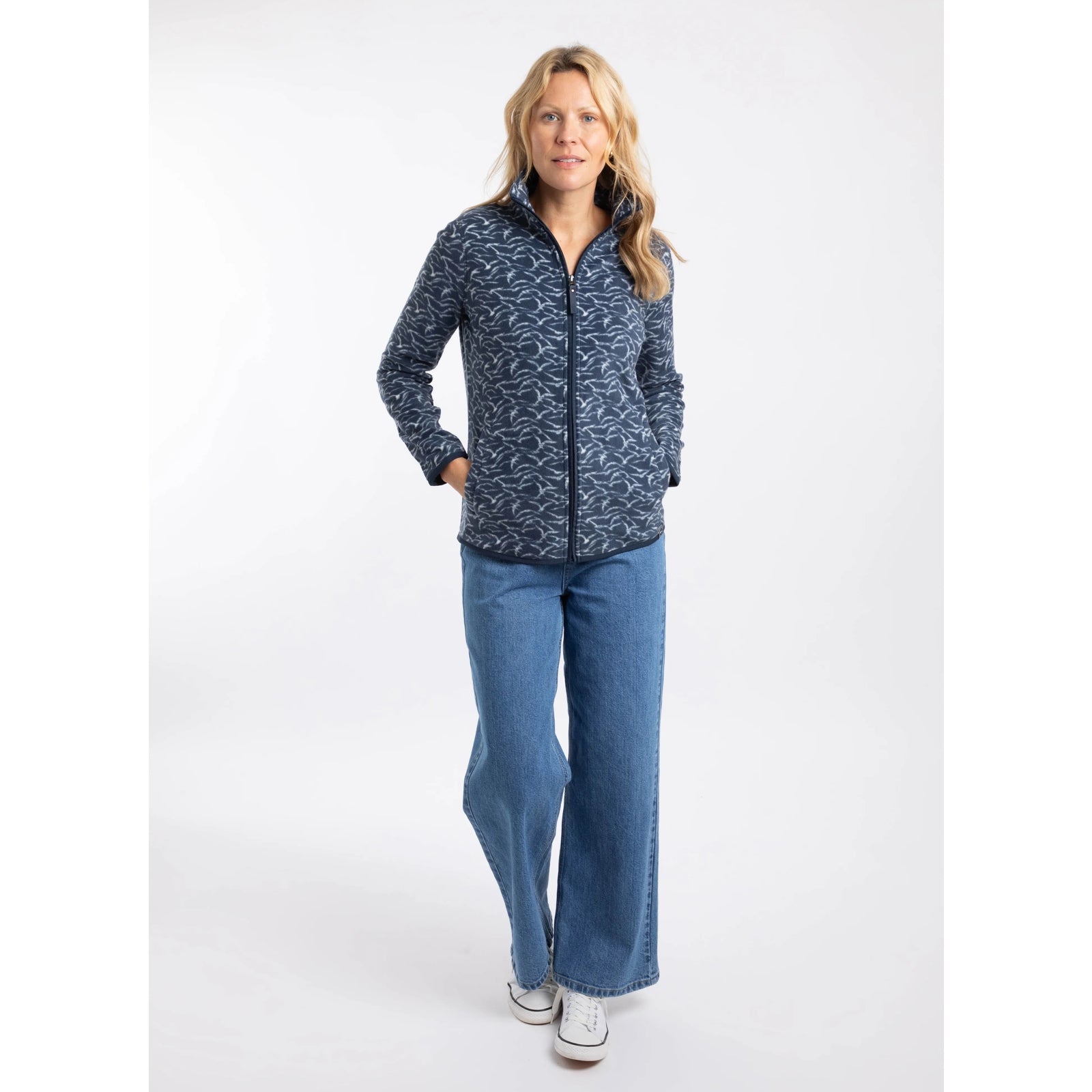 A woman wearing a Weird Fish full zip Rochelle microfleece in Night Blue with a gull bird print with blue jeans and white trainers.
