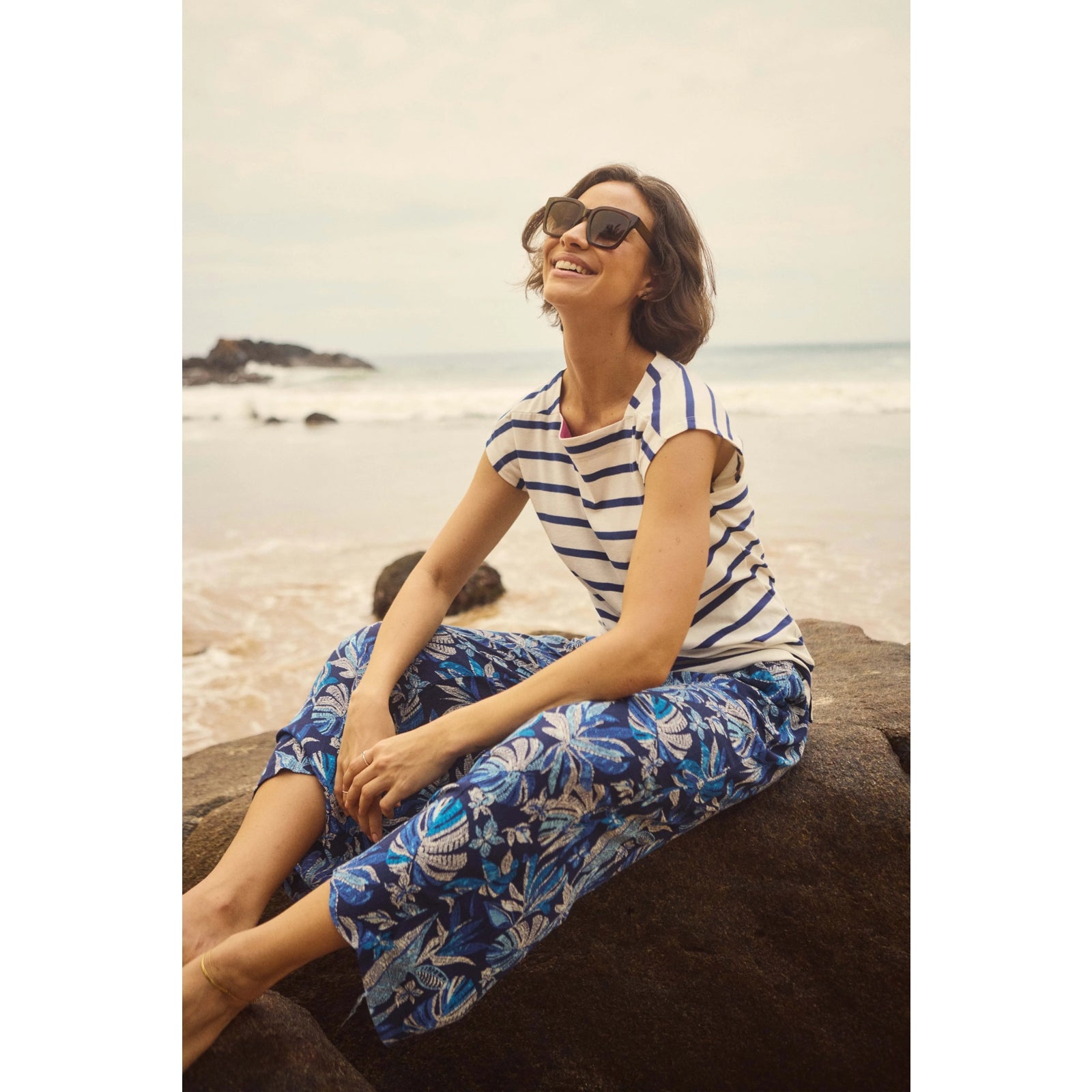 A woman sat by the coast wearing a pair of Weird Fish Sarita cropped trousers in indigo blue with a tropical leaf pattern.