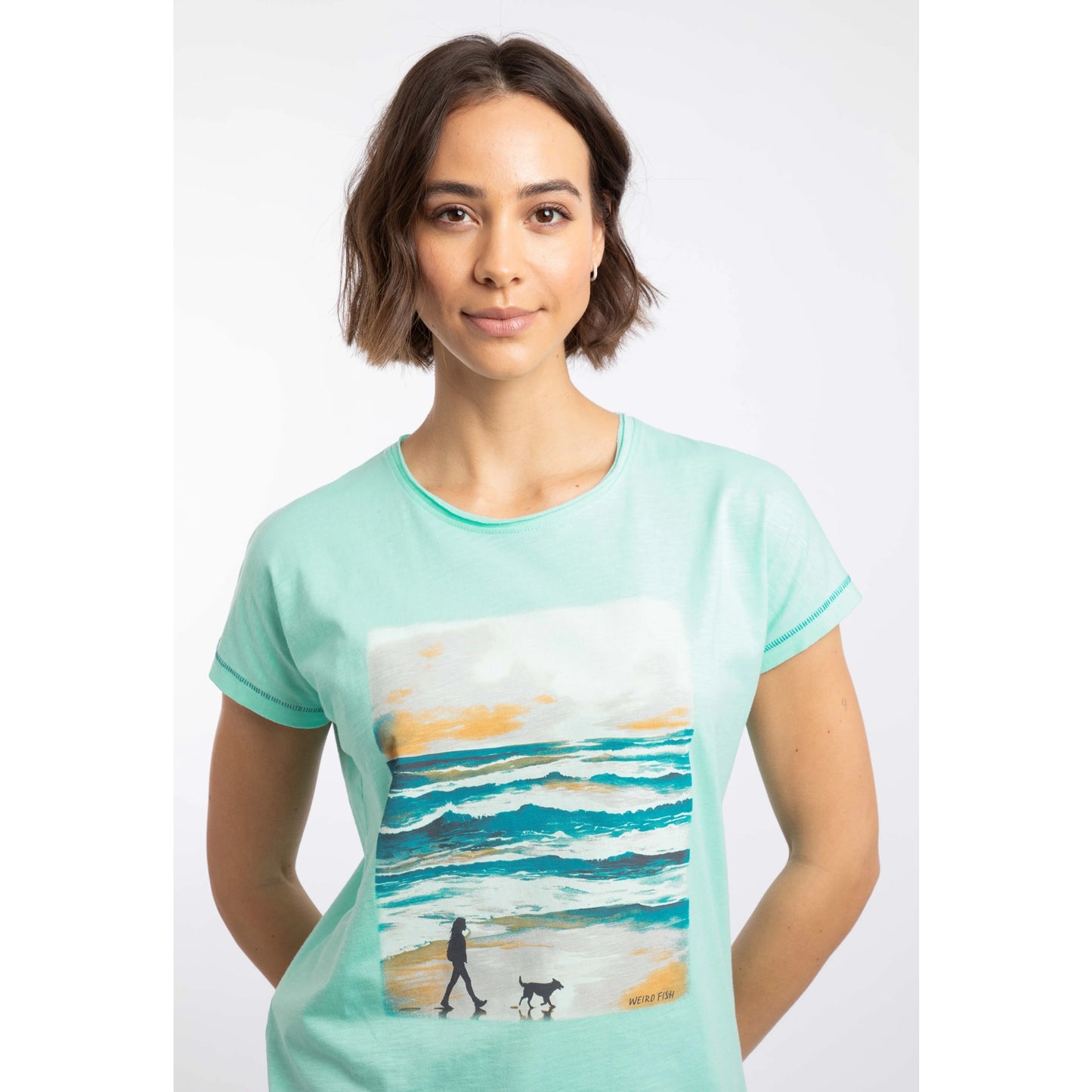 Woman wearing a Weird Fish Apple Mint colour t-shirt with a dog walking beach scene design on a white background
